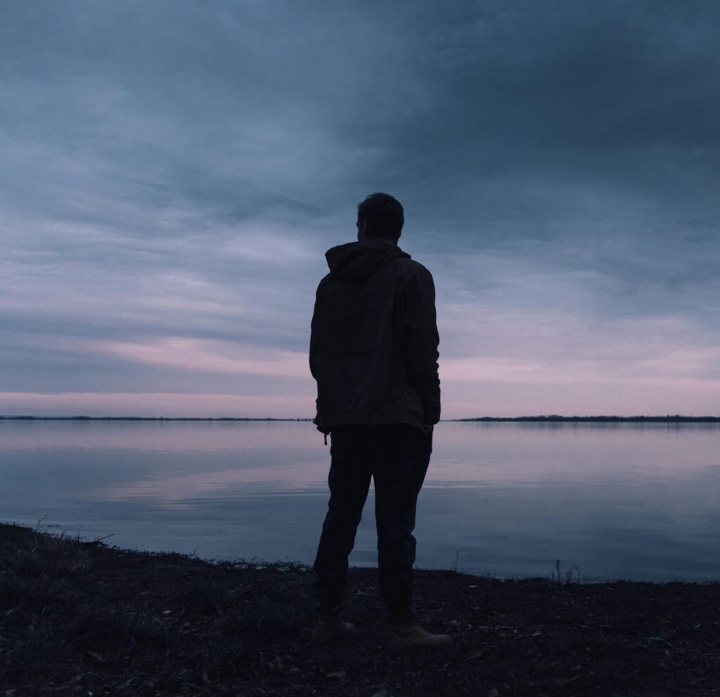 A solitary silhouette of a man in a jacket gazing at a lake during a peaceful sunset, creating a serene atmosphere.